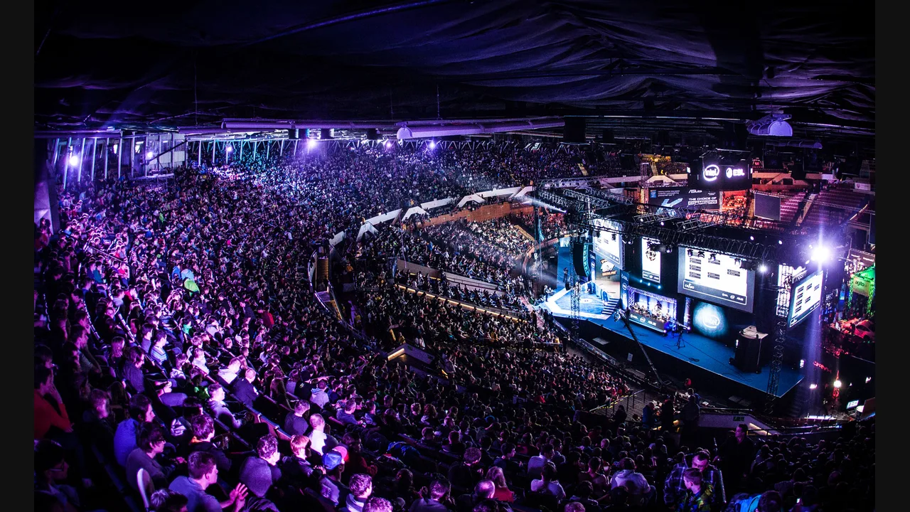 Photograph of a packed esports arena crowd facing a large stage with glowing screens